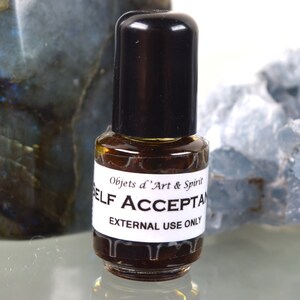 May include: A small brown glass bottle with a black cap. The bottle is labelled "Objets d'Art & Spirit" and "SELF ACCEPTANCE" with the text "EXTERNAL USE ONLY" below. The bottle is sitting on a white surface.