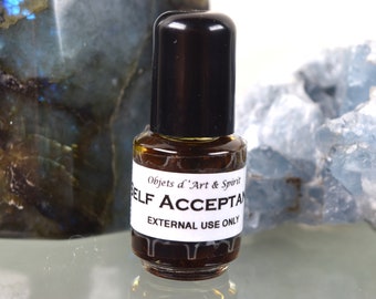 May include: A small brown glass bottle with a black cap. The bottle is labeled "Objets d'Art & Spirit" and "SELF ACCEPTANCE" with the text "EXTERNAL USE ONLY" below. The bottle is sitting on a white surface.