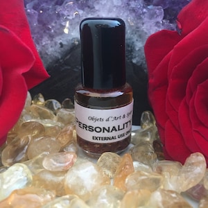 May include: A small glass bottle with a black cap, filled with a brown liquid. The label reads "Personality" and "Objets d'Art & Spirit." The bottle is surrounded by small, light-colored stones and red roses.