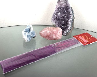 May include: A package of purple incense sticks with a red label that says "Gardenia" and "Natural Incense". The package is sitting on a glass surface with three crystals: a pink rose quartz, a light blue crystal, and a purple amethyst.