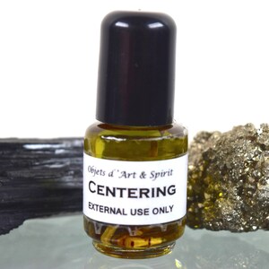 May include: A small glass bottle with a black cap, filled with a yellow liquid, labeled "CENTERING" and "EXTERNAL USE ONLY." The bottle is flanked by a black tourmaline crystal and a gold-colored pyrite stone.