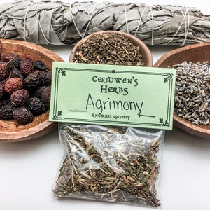 May include: A green label with the text "Ceridwen's Herbs Agrimony External Use Only" is attached to a clear plastic bag filled with dried herbs. The bag is surrounded by three wooden bowls, each containing different dried herbs. One bowl contains dried rose hips, another contains dried lavender, and the third contains dried herbs.