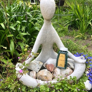 May include: A white concrete sculpture of a person sitting in a meditative pose with a small green glass bottle of essential oil in their lap. The bottle has a gold label with the text "Pure Essential Oil" and "Peppermint" on it. The sculpture is surrounded by green plants and rocks.