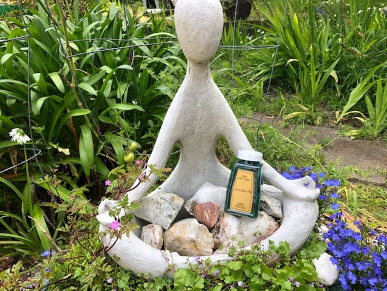 May include: A white concrete sculpture of a person sitting in a meditative pose with a small green glass bottle of essential oil in their lap. The bottle has a gold label with the text "Pure Essential Oil" and "Peppermint" on it. The sculpture is surrounded by green plants and rocks.