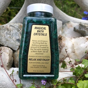 May include: A green plastic bottle with a white lid containing bath crystals. The label reads "Magical Bath Crystals" and includes instructions for use. The bottle is sitting on a bed of rocks and green foliage.