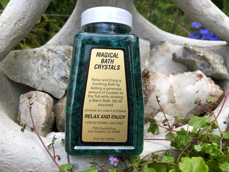 May include: A green plastic bottle with a white lid containing bath crystals. The label reads "Magical Bath Crystals" and includes instructions for use. The bottle is sitting on a bed of rocks and green foliage.