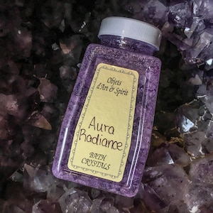 Peut inclure: Une bouteille en plastique transparente remplie de cristaux de bain violets. La bouteille a un couvercle blanc et une étiquette jaune sur laquelle est inscrit "Aura Radiance Bath Crystals" et "Objets d'Art & Spirit".