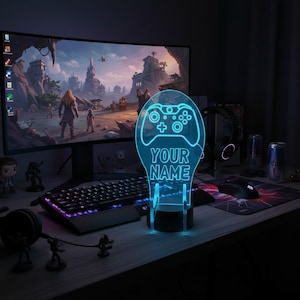 May include: A custom LED light sign featuring a glowing cyan outline of a video game controller and the text "YOUR NAME" etched onto a clear acrylic panel, supported by a black circular base, positioned on a desk in a gaming setup.