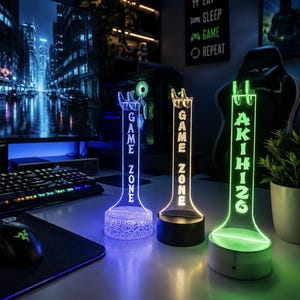 May include: Three vertical acrylic display stands with illuminated bases. Two stands feature the text GAME ZONE in white and yellow light, while the third displays the name AKIHI26 in green light. Each stand has a notched top for holding items.