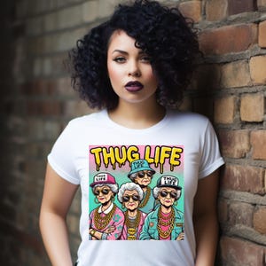 Puede incluir: Camiseta blanca con un gráfico colorido de cuatro mujeres mayores con gafas de sol, cadenas de oro y sombreros, con las palabras "THUG LIFE" en amarillo. Las mujeres están representadas en un estilo de dibujos animados.