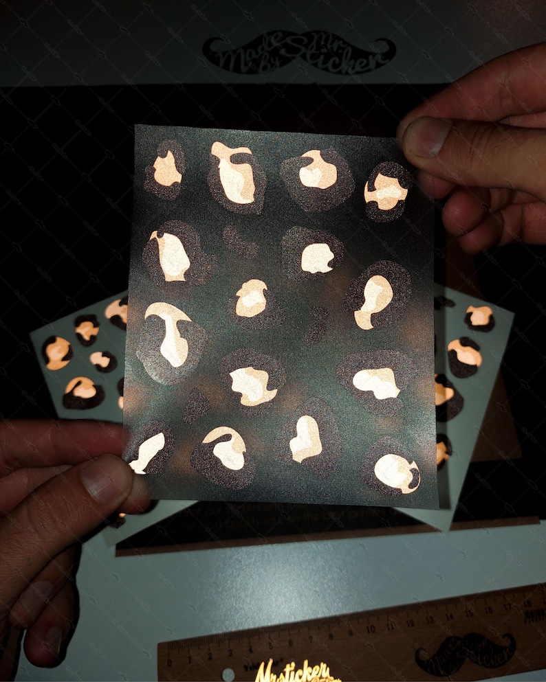 May include: A sheet of clear vinyl with a gold glitter leopard print design. The sheet is being held by two hands. The text "Made by Mr. Sticker" is visible in the background.
