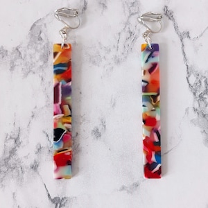 Op de afbeelding: Een paar kleurrijke, lange, rechthoekige clip-on oorbellen met een veelkleurig marmerpatroon. De oorbellen zijn gemaakt van acryl en hebben zilverkleurige metalen clips.