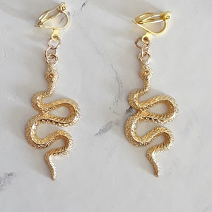 Peut inclure: Une paire de boucles d'oreilles clips en forme de serpent dorées. Les serpents sont enroulés et ont une surface texturée.