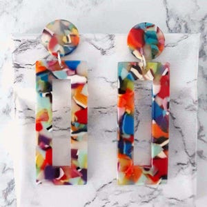 Peut inclure: Une paire de boucles d'oreilles géométriques colorées. Chaque boucle d'oreille est composée d'un clou circulaire et d'une goutte rectangulaire avec un centre évidé. Les boucles d'oreilles présentent un motif mosaïque aux couleurs vives.