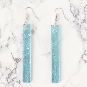 Peut inclure: Une paire de longues boucles d'oreilles pendantes bleu clair avec un motif floral. Les boucles d'oreilles sont en verre et ont des crochets argentés.