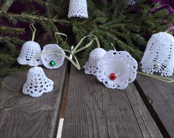 Op de afbeelding: Witte gehaakte bellen met groene en rode belletjes die aan een dennentak hangen op een houten oppervlak.