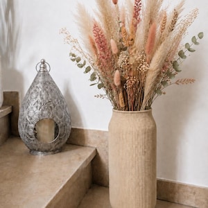 Könnte beinhalten: Eine hohe, zylindrische beige Vase mit gerippter Textur enthält ein großes Arrangement aus getrocknetem Pampasgras, rosa Hasenschwänzchen, Weizenähren und Eukalyptuszweigen. Daneben steht eine silberfarbene Metalllaterne mit Ausschnitten.