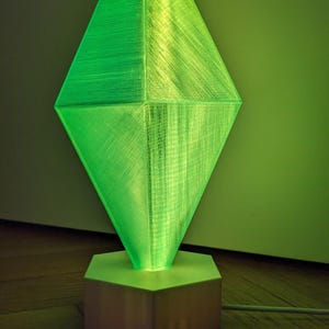 Puede incluir: Una lámpara verde en forma de diamante con una base hexagonal blanca. La lámpara está encendida y proyecta un brillo verde.
