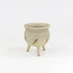 May include: A small, round, three-legged ceramic planter with a speckled, light beige and muted green glaze. The vessel features several diagonal, almond-shaped cutouts along its midsection, providing ventilation and a distinct, open design.