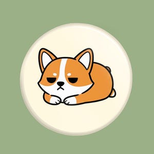 May include: A round, cream-colored button featuring a cartoon corgi dog with orange and white fur, and a grumpy expression. The dog is lying down with its paws crossed. The background is a solid, muted green.