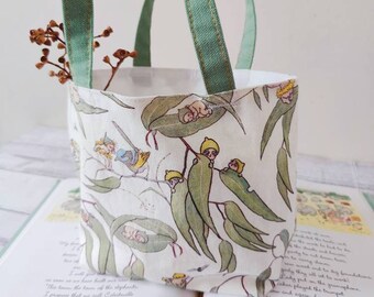 May include: A small green canvas tote bag with a white fabric lining featuring a repeating pattern of small green leaves and tiny fairies. The bag has two handles and is sitting on top of an open book.