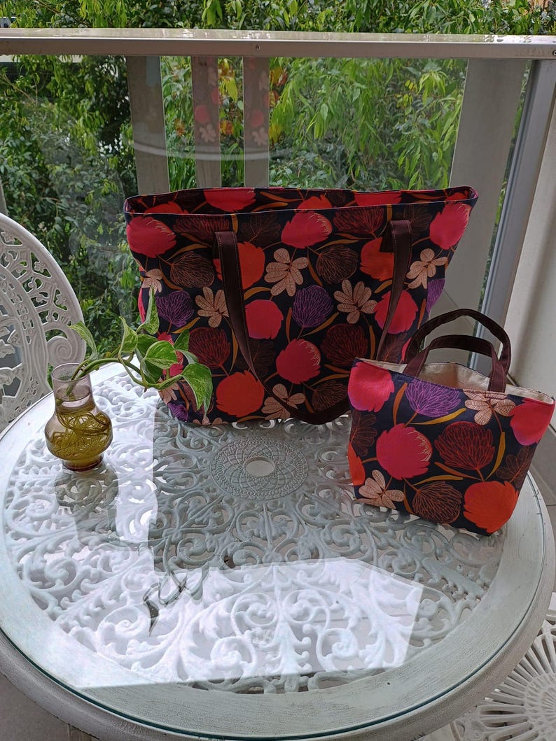 May include: Two tote bags with floral patterns. The larger bag is black with pink, red, and purple flowers. The smaller bag is a similar pattern but in a smaller size.