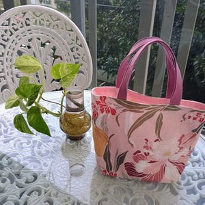 May include: A pink tote bag with a floral pattern featuring red and white flowers and brown leaves. The bag has magenta handles and a pink interior. The bag is sitting on a glass table with a white decorative chair and a small plant in the background.