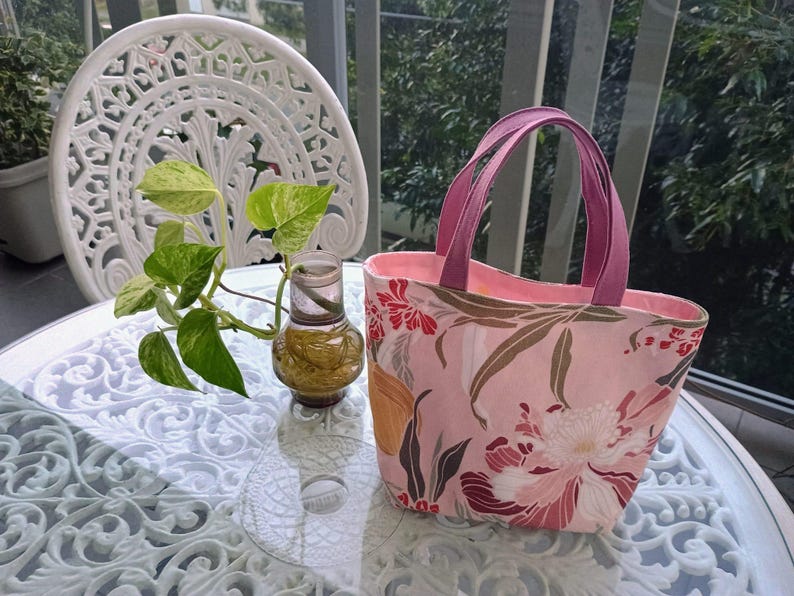May include: A pink tote bag with a floral pattern featuring red and white flowers and brown leaves. The bag has magenta handles and a pink interior. The bag is sitting on a glass table with a white decorative chair and a small plant in the background.