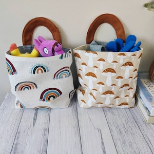 Op de afbeelding: Twee canvas opbergmanden met bruine leren handvatten. De ene mand heeft een regenboogpatroon en de andere een paddenstoelpatroon.