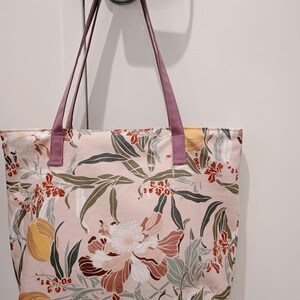 May include: A pink tote bag with a floral pattern featuring large flowers, leaves, and fruit in shades of red, brown, yellow, and green. The bag has purple handles and is made of fabric.