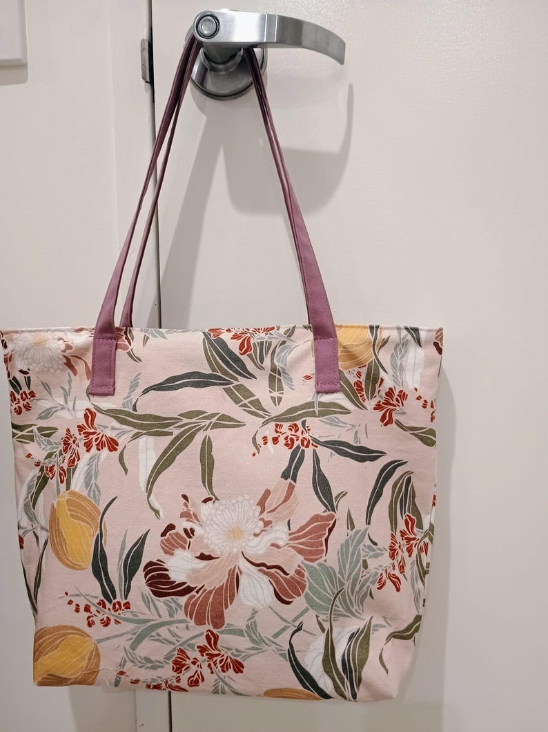 May include: A pink tote bag with a floral pattern featuring large flowers, leaves, and fruit in shades of red, brown, yellow, and green. The bag has purple handles and is made of fabric.