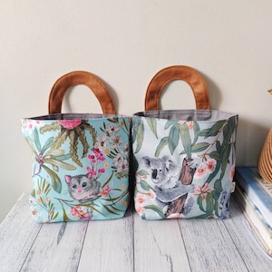 May include: Two fabric storage bags with brown handles. The bag on the left has a blue background with a floral pattern and a possum. The bag on the right has a blue background with a floral pattern and a koala.