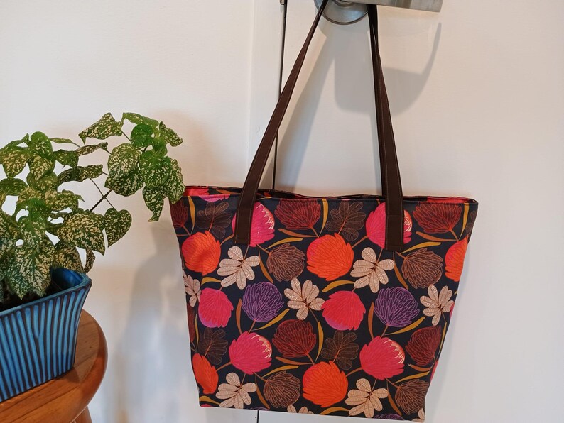 May include: A brown tote bag with a floral print featuring pink, orange, and purple flowers on a dark background. The bag has brown straps.
