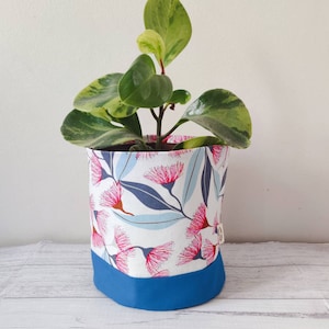 May include: A white fabric planter with a blue trim and a pink floral pattern. The planter is filled with a green plant with variegated leaves.