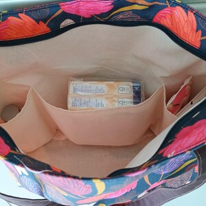 May include: A black tote bag with a floral print featuring pink, orange, and yellow flowers. The bag has a zipper closure and multiple interior pockets. The bag is lined with a beige fabric.