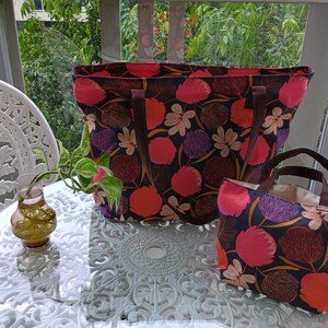 May include: Two tote bags with floral patterns. The larger bag is black with pink, red, and brown flowers. The smaller bag is black with pink, red, and gold flowers. Both bags have brown leather handles.