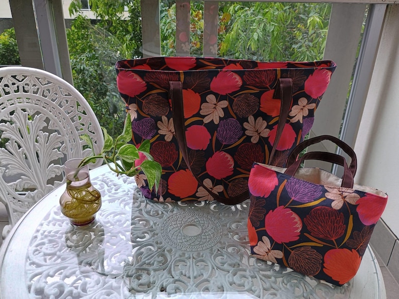 May include: Two tote bags with floral patterns. The larger bag is black with pink, red, and brown flowers. The smaller bag is black with pink, red, and gold flowers. Both bags have brown leather handles.
