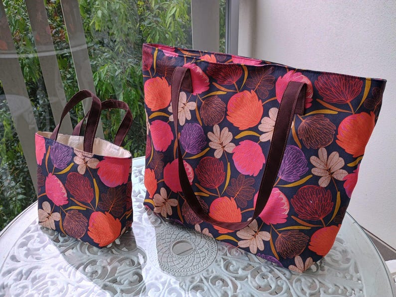 May include: Two tote bags with floral patterns. The larger bag is navy blue with pink, orange, and purple flowers. The smaller bag is red with pink, orange, and purple flowers.