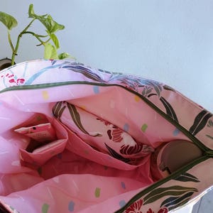 May include: Open tote bag with a floral pattern in pink, green, and white. The interior is pink with multiple pockets and a zippered closure. A small plant is visible in the background.