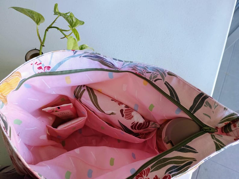 May include: Open tote bag with a floral pattern in pink, green, and white. The interior is pink with multiple pockets and a zippered closure. A small plant is visible in the background.