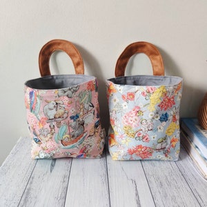 May include: Two fabric storage bins with brown handles. The bin on the left has a pink background with a colourful pattern of Australian animals and plants. The bin on the right has a light blue background with a colourful pattern of flowers and plants.