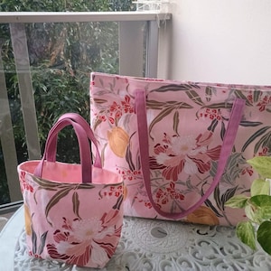 May include: Two pink floral tote bags. The larger bag has long purple handles, and the smaller bag has short purple handles. The bags feature a repeating pattern of flowers, leaves, and fruit in shades of pink, red, and brown.