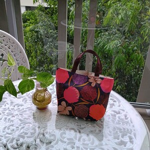May include: A brown tote bag with a floral print featuring pink, red, and purple flowers on a dark background. The bag has brown handles and is sitting on a white table.