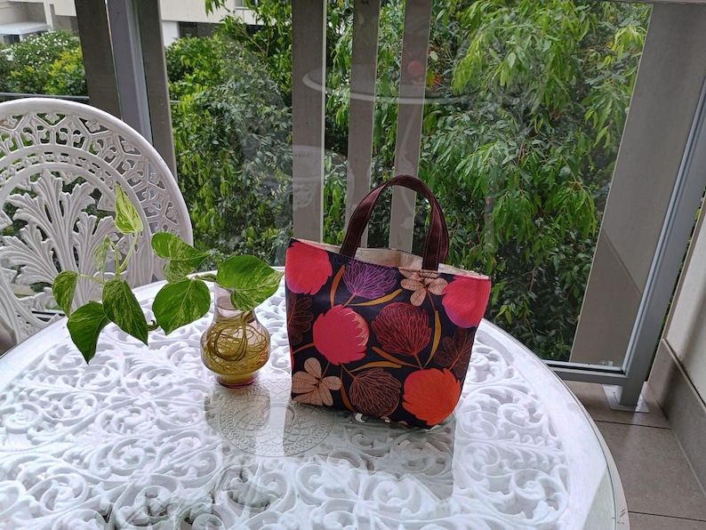 May include: A brown tote bag with a floral print featuring pink, red, and purple flowers on a dark background. The bag has brown handles and is sitting on a white table.