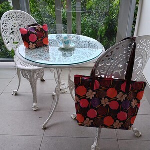 May include: Two white wrought iron chairs and a table with a glass top on a balcony. A floral patterned tote bag is hanging on the back of one of the chairs. A floral patterned handbag is on the table. A cup and saucer are on the table.