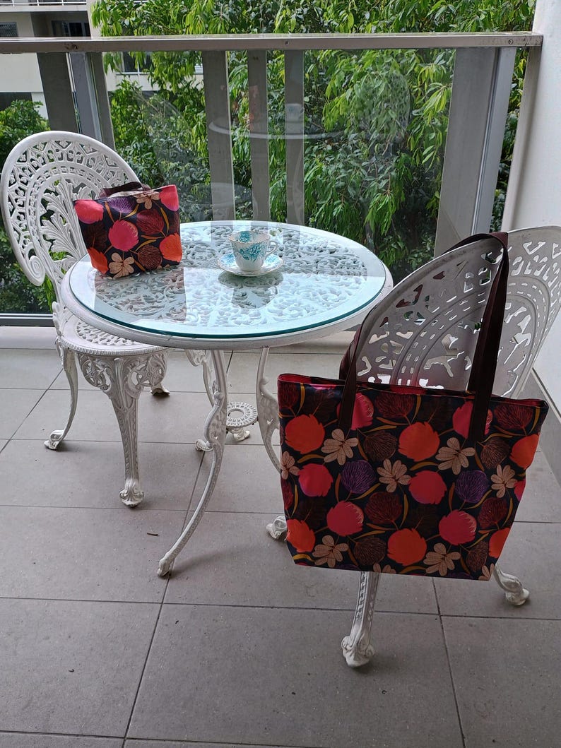 May include: Two white wrought iron chairs and a table with a glass top on a balcony. A floral patterned tote bag is hanging on the back of one of the chairs. A floral patterned handbag is on the table. A cup and saucer are on the table.
