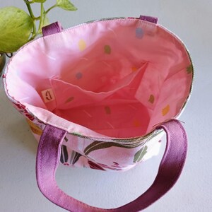 May include: Open view of a small tote bag with a floral pattern. The exterior features a pink and white floral design with purple handles. The interior is a solid pink with small pockets. The bag is designed for carrying small items.