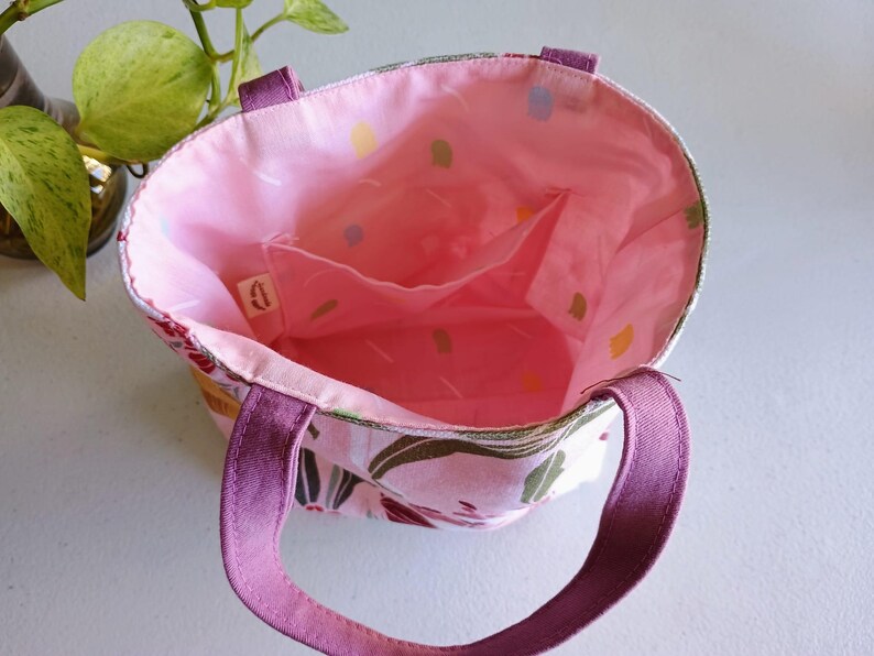 May include: Open view of a small tote bag with a floral pattern. The exterior features a pink and white floral design with purple handles. The interior is a solid pink with small pockets. The bag is designed for carrying small items.