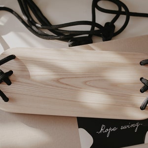 May include: A wooden swing set with black rope and metal clips. The swing is made of natural wood and has a simple, minimalist design. The rope is tied to the swing with a knot and the metal clips are used to attach the rope to the swing.
