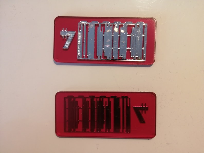 May include: Two rectangular, red-tinted plastic pieces. The top piece has silver-colored shapes and the number 7. The bottom piece has black shapes and the number 7. The shapes appear to be components or parts.